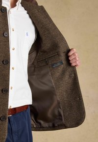 Brown herringbone tweed jacket with a button closure, featuring a smooth brown satin lining and a label sewn into the interior pocket.