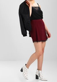 Top nero senza maniche con pizzo e dettagli in bottoni, gonna borgogna con bordo in pizzo e stivaletti bianchi con borchie. Giacca nera in mano.