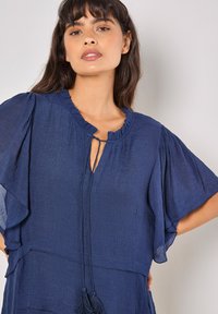 Marineblaue Bluse mit flatternden Ärmeln, strukturiertem Stoff, Rüschen-Halsausschnitt und Kordelzugverschluss mit Quasten.