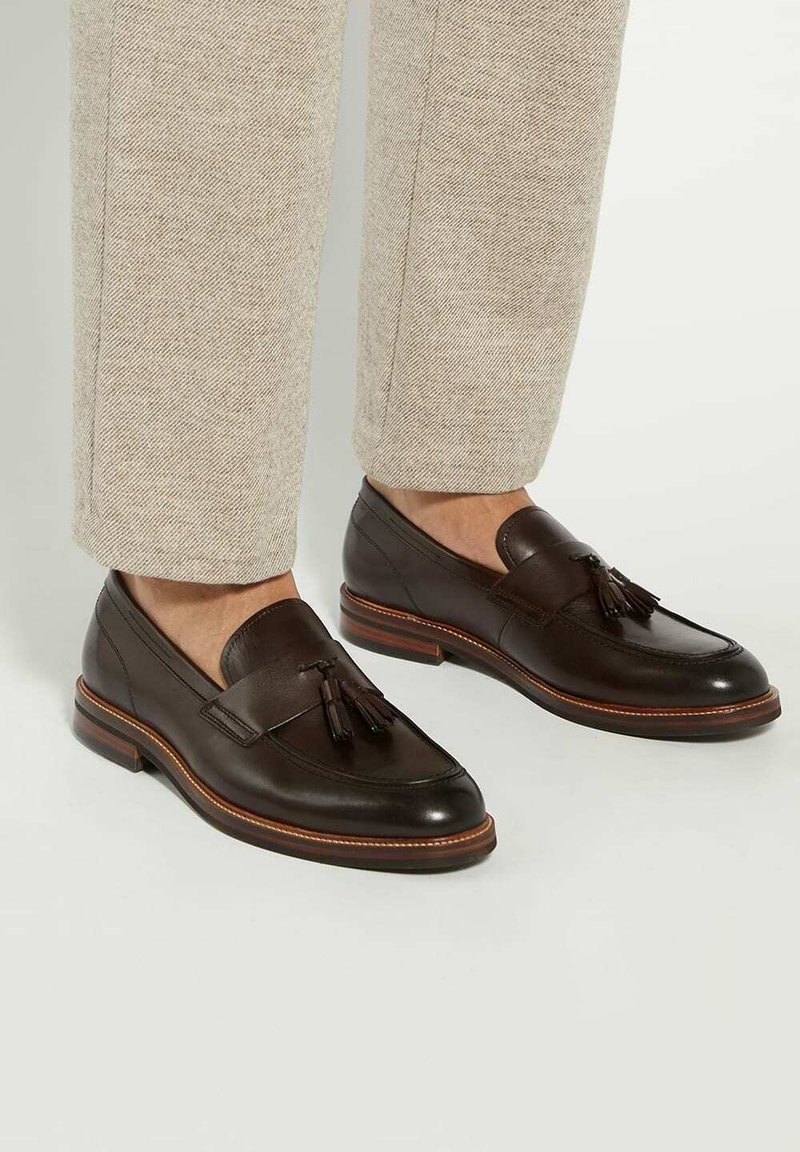 Brown leather loafers with tassel detail, smooth finish, and contrasting lighter stitching. Pairs with beige textured trousers.