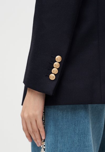 Μανίκι μπλε σακακιού με τέσσερα χρυσά ανάγλυφα κουμπιά πάνω από μπλε τζιν, χέρι χαλαρό στο πλάι κρατώντας ένα κόσμημα με χάντρες.