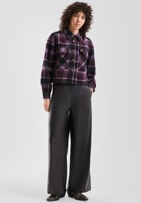 Przycięta czarno-fioletowa koszula flanelowa w kratę z dwoma kieszeniami na piersi i zapięciem na guziki, w zestawieniu z czarnymi szerokimi spodniami i butami.