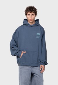 Sudadera con capucha en algodón azul con un bolsillo frontal y corte holgado. Presenta texto impreso en el pecho. Textura suave, diseño casual.