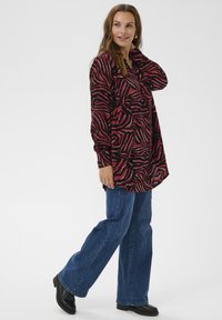 Long-sleeve shirt in red and black swirl pattern, paired with blue flared jeans and black boots. Fabric appears soft with a relaxed fit.