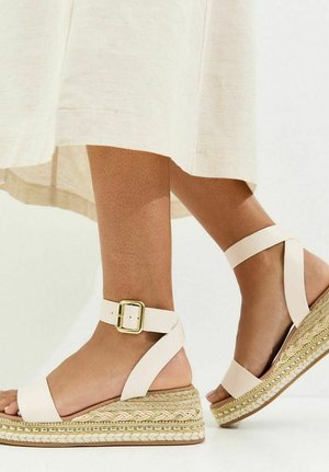 Voeten met beige sleehakken met enkelbandjes en gouden gespen, gecombineerd met een lichtgekleurde midirok.