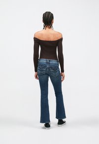 Osoba s pletenými vlasmi, oblečená v hnedom topu s odhalenými ramenami, modrých rozšírených džínsoch a čiernych teniskách, stojaca chrbtom k bielej stene.