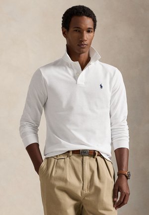 Uomo che indossa una polo bianca a maniche lunghe con logo blu, pantaloni beige, cintura marrone e orologio da polso marrone su sfondo neutro.