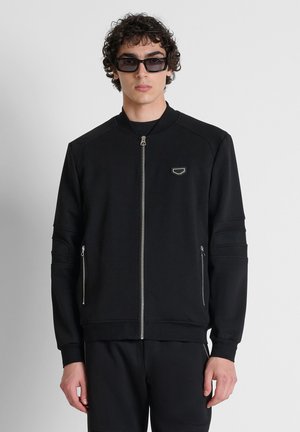 Young man with curly hair wearing black zip-up jacket, matching pants, and dark rectangular sunglasses, standing against plain background.