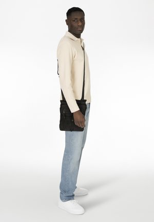 Borsa a tracolla in suede nero con tasche con zip, indossata sulla spalla. Modello con una maglietta beige chiara e jeans azzurri chiari in piedi su uno sfondo bianco.