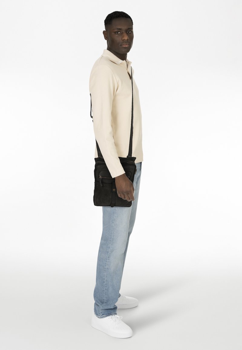 Bolso cruzado de ante negro con bolsillos con cremallera, llevado al hombro. Modelo con una blusa beige clara y jeans azules claros, de pie frente a un fondo blanco.