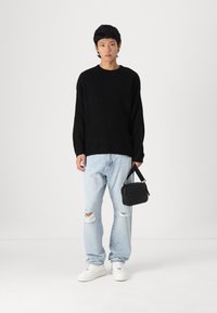Pull en maille côtelée noir, jean déchiré bleu clair, baskets blanches, sac à main noir avec bandoulière, se tenant devant un fond uni.