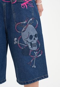 Denim shorts with a relaxed fit, featuring a skull design embellished with rhinestones, pink accents, and contrasting stitching.