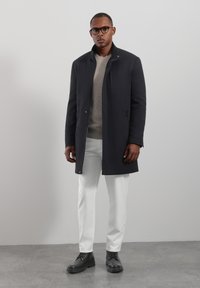 Donkergrijze jas met een gestructureerde kraag, knoopsluiting aan de voorkant, twee zijzakken; gecombineerd met een lichtgrijze trui, witte broek en zwarte laarzen.