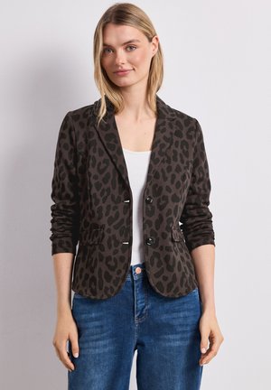 Junge Frau trägt einen dunklen Blazer mit Leopardenmuster über einem weißen Top und blauen Jeans, stehend vor einem hellgrauen Hintergrund.