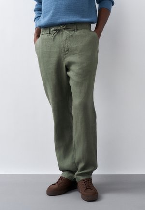 Άνδρας που φοράει χακί παντελόνι από λινό με ελαστική μέση με κορδόνι, τα χέρια στις τσέπες, καφέ αθλητικά παπούτσια και γαλάζιο πουλόβερ.