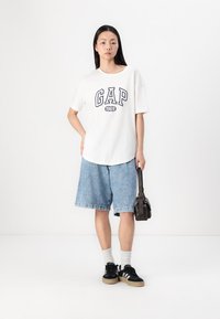 Balta īsām piedurknēm T-kreklā ar zilu "GAP 1969" logo, zili džinsa šorti, balti apkalpju zeķes, melni sporta apavi un neliela melna crossbody soma.