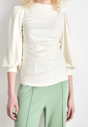 Kvinde iført en cremefarvet langærmet rynket bluse med puffede ærmer og lysegrønne højtaljede bukser, stående mod en ensfarvet baggrund.