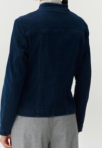 Kvinna som bär en mörkblå denimjacka och ljusgrå byxor, sedd bakifrån mot en enkel ljus bakgrund.