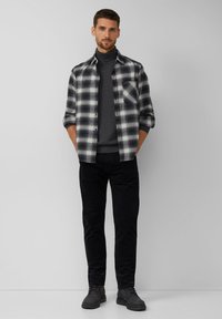 Koszula w kratę w czerni i bieli, noszona na szarym golfie, zestawiona z czarnymi spodniami i ciemnymi butami, stojąca na neutralnym tle.