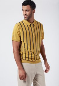 Gul randig pikétröja med zipkrage och korta ärmar, med vertikala marinblå ränder. Bärs med beige byxor.