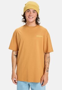 Kortærmet, sennepsfarvet t-shirt med rund halsudskæring og lille logo. Bæret sammen med lyseblå jeans, modellen har tatoveringer og en beanie.
