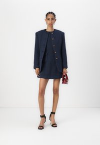 Tumesinine tweed ülikond, millel on struktureeritud jakki ja mini seelik, nelja kuldne nööbi ja ees olevate taskutega. Mustad rihmadega kontsad ja punane käekott.