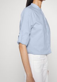 Kvinna klädd i en ljusblå, långärmad skjorta med uppvikta ärmar och vita byxor, tagen från sidan mot en enkel bakgrund.