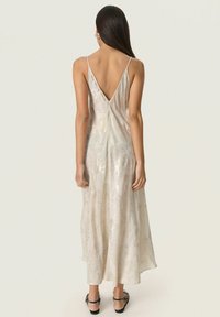 Woman wearing a sleeveless, shimmering cream midi dress with a deep V-back and black open-toe heels standing against a plain light background.