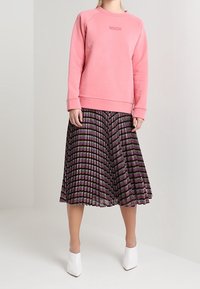 Sudadera rosa con mangas raglán que presenta el logo "MSCH", combinada con una falda plisada de longitud a la rodilla en franjas negras, moradas y verdes.