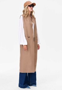 Beżowa, bezrękawnikowa kamizelka z miękkiego materiału, z dwiema przednimi kieszeniami. Noszona na białą bluzkę i zestawiona z rozkloszowanymi, ciemnymi dżinsami.