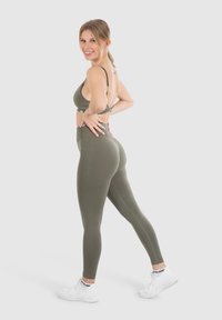 Reggiseno sportivo verde oliva e leggings a vita alta realizzati in tessuto elastico. Le sneakers sono bianche con dettagli colorati. La modella posa con le mani sui fianchi.