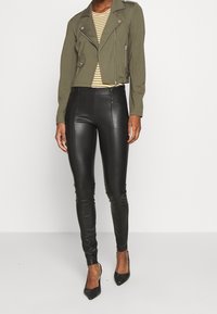 Person wearing black faux leather leggings, olive green cropped jacket, striped yellow top, and black pointed heels standing against white background.