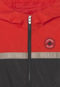 Rød og sort vindjakke med lynlås og et rundt logo patch. Refleksbånd med "CONVERSE" langs taljen, letvægtsmateriale.