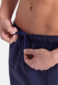 Pantaloni da bagno blu navy con vita elastica e cordoncino. Il tessuto è liscio con una texture leggera. Primo piano di mani che regolano il fiocco.