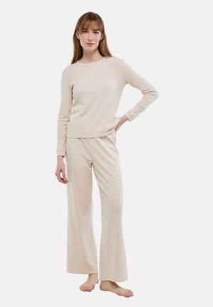 Femme debout pieds nus portant un haut beige à manches longues et un pantalon large avec des détails de boutons sur le haut et la ceinture.