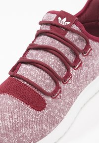 Mörkröd träningssko med en strukturerad stickad ovandel, elastiska snören och en vit sula. Har en logotypflik på tungan och en vadderad krage.