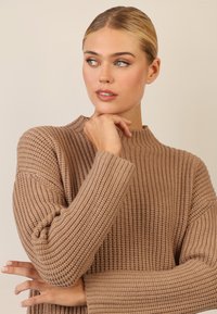 Taupe gerippter Strickpullover mit hohem Kragen und lockeren Ärmeln, der eine strukturierte Oberfläche und einen lässigen Schnitt aufweist.