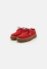 Zapatos de ante rojo con punta redonda, suela plana de goma marrón y cordones. Presentan costuras a lo largo de los bordes y una suela texturizada.