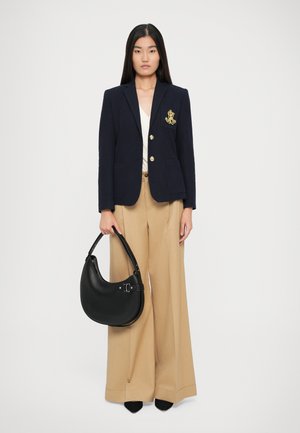Blazer marine avec boutons dorés et emblème, associé à un pantalon ample beige. Le modèle tient un sac hobo en cuir noir. Textures de tissu lisses.