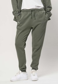 Persona che indossa pantaloni della tuta verde oliva con coulisse, sneaker bianche e una giacca abbinata verde oliva con le mani nelle tasche.