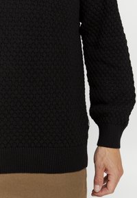 Svart teksturert genser med strikket mønster og ribbede mansjetter. Har et lite logomerke på siden og en avslappet, tilpasset silhuett.