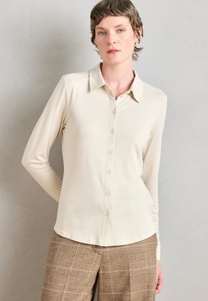 Beige long-sleeve shirt with a collared neckline, featuring a button-up front and smooth texture, paired with plaid, brown trousers.