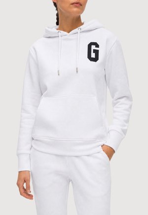 Femme portant un sweat à capuche blanc avec une lettre G noire sur le côté gauche de la poitrine et un pantalon blanc assorti, debout avec une main dans la poche.