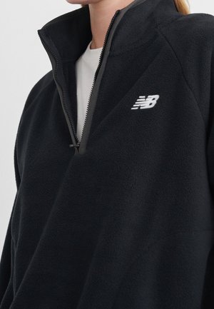 Person iført en sort New Balance fleecejakke med en halv lynlås over en hvid skjorte, hvor kun den øverste del af brystet og halsen vises.