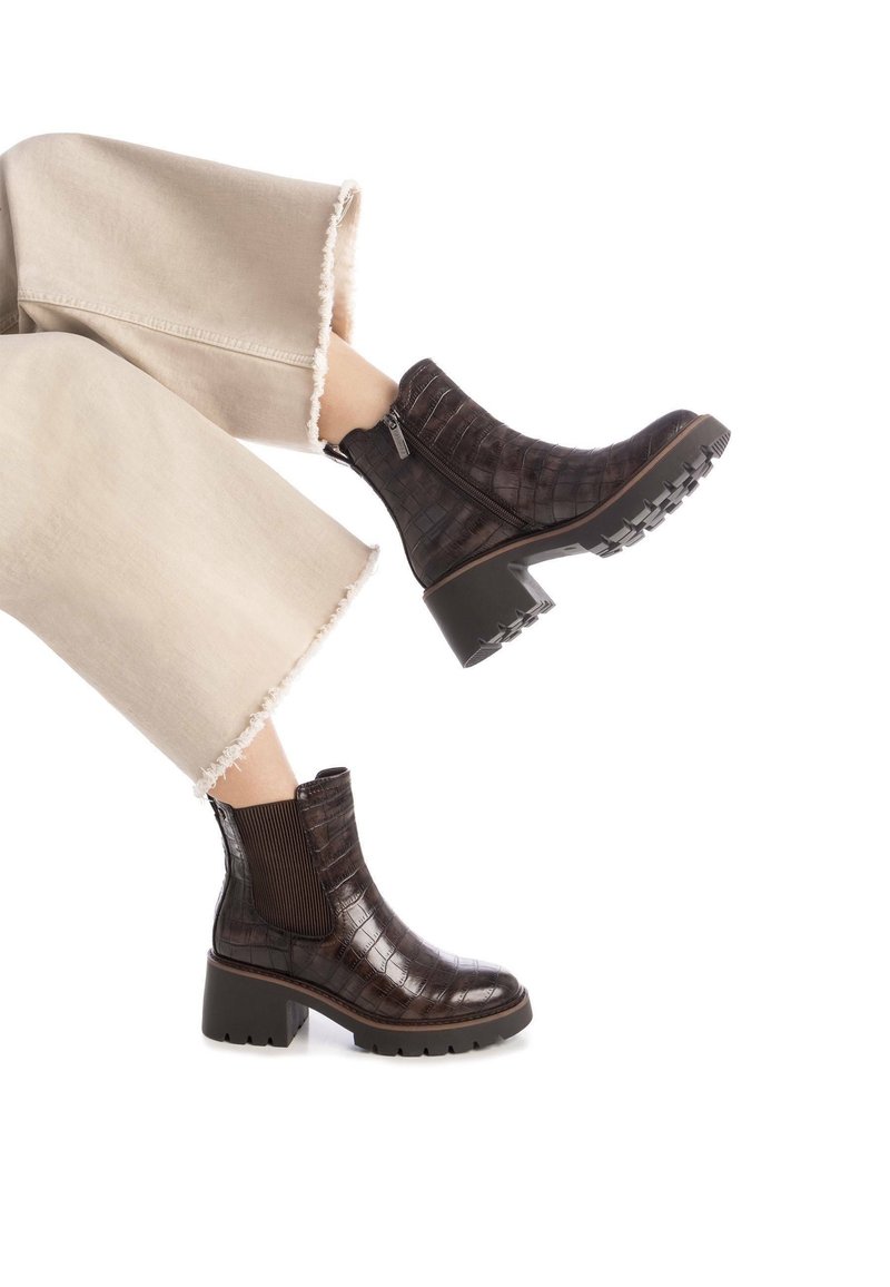 Vue en longueur des jambes d'une personne portant un pantalon large beige effiloché et des bottines en cuir embossé crocodile marron foncé à talons blocs.