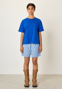 Mėlyna medvilnė marškinėliai su trumpomis rankovėmis, derinami su mėlynomis ir baltomis dryžuotomis šortais bei rudais iki kelių aukštakulniais batais su sagtimis.