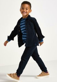 Marineblå kordfløjlsskjorte, blå stribet skjorte indenunder, marineblå bukser, brune velcro-sneakers. Glat tekstur, afslappet pasform og lagdelt design.