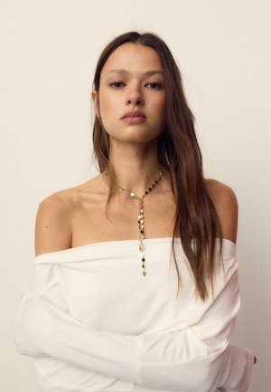 Žena s dugom smeđom kosom nosi bijelu majicu bez ramena i zlatnu naušnicu u obliku geometrijskog privjeska na jednostavnoj pozadini.