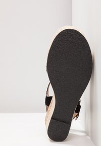 Sandale compensée avec semelle en caoutchouc noire texturée, semelle intermédiaire beige tressée et brides noires, positionnée semelle vers le haut sur fond blanc.