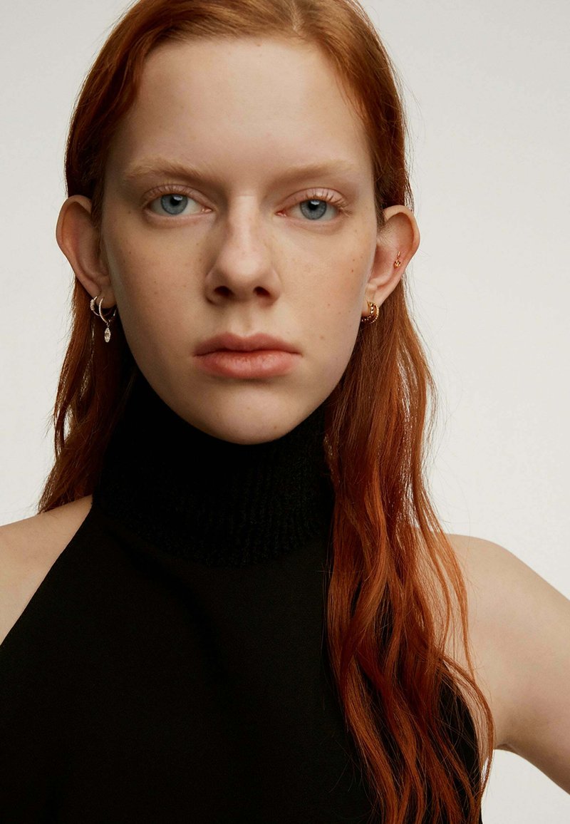 Top negro sin mangas con un cuello alto y acanalado. La modelo tiene el cabello largo y ondulado de color rojo y lleva pendientes pequeños en forma de aro con un detalle colgante.